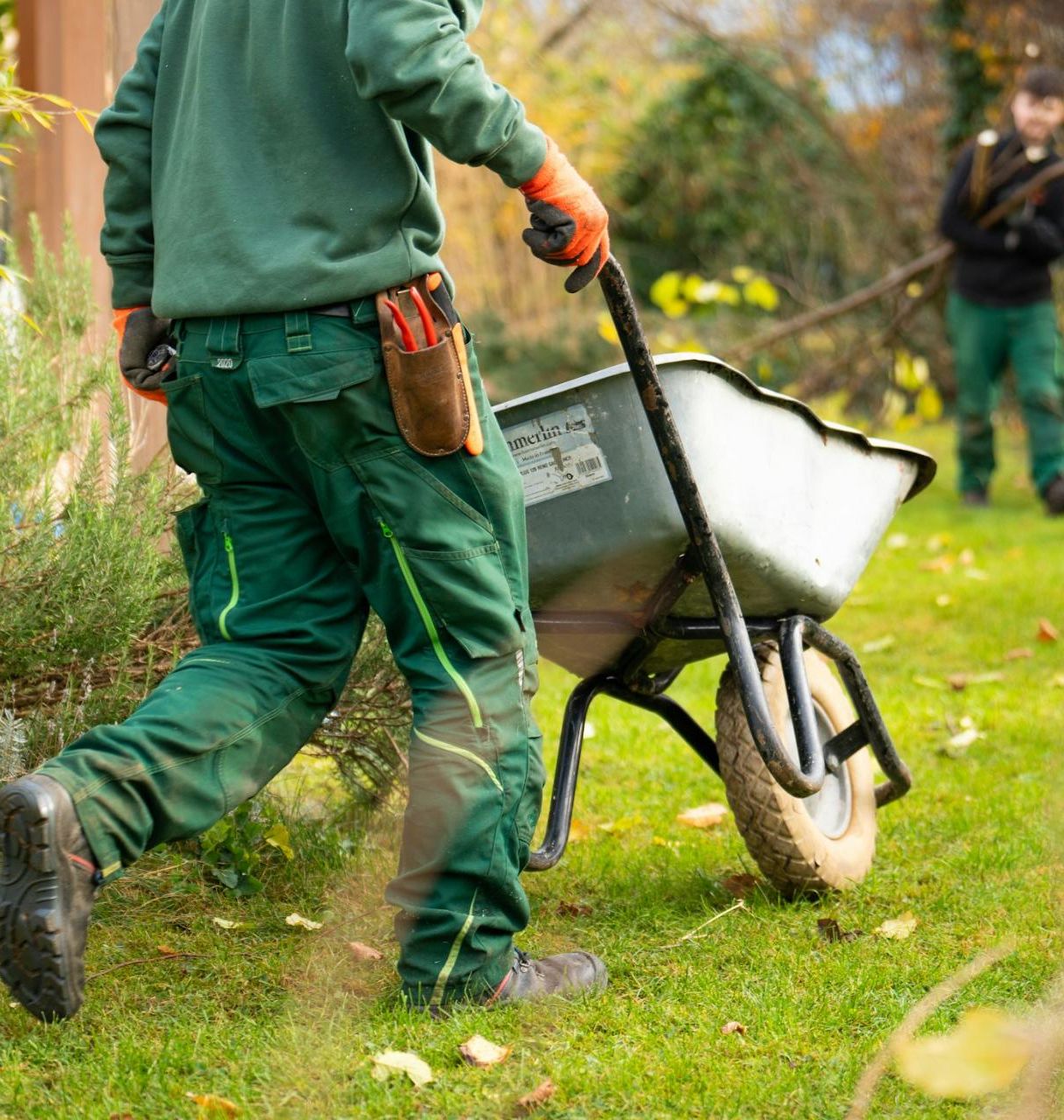 Two landscape crew members in uniform working on a property, one pushing a wheelbarrow